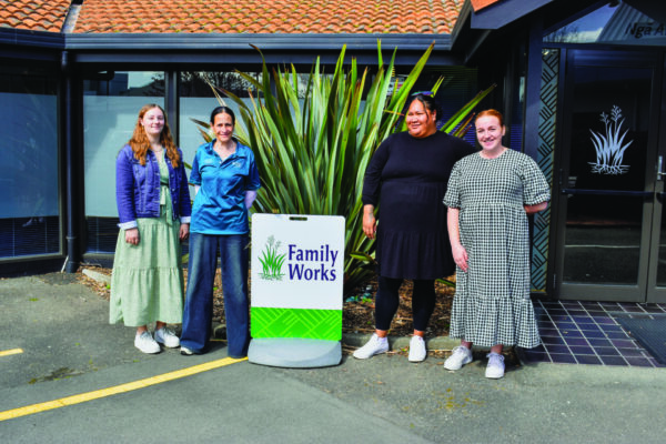 Some of the dedicated Family Works team supporting through our local schools. Four team members are standing outside the Family Works office in Hastings with a sign that says Family Works. There is a harakeke plant behind them.