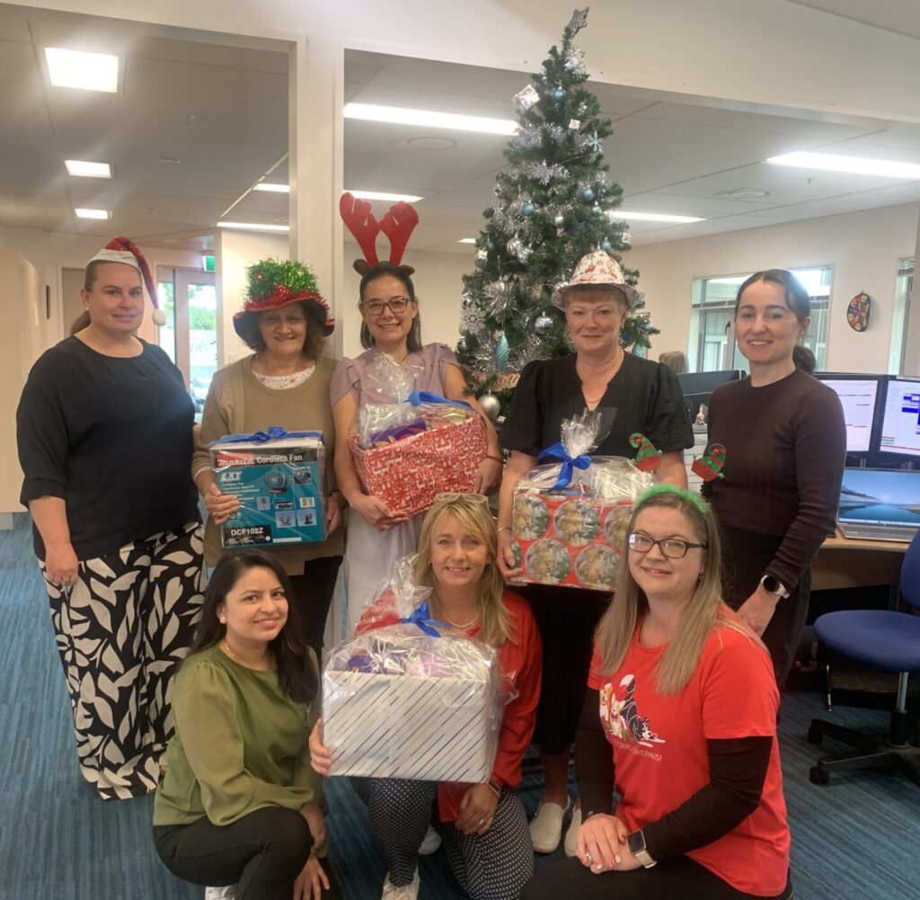 Eight of the Enliven Older People team stand in an office holding four raffle boxes. There is a Christmas tree behind them. The team are wearing Christmas hats and decorations and are smiling.